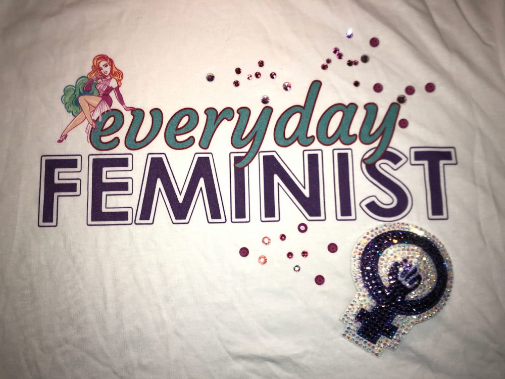 Photo of a shirt that says everyday feminist, with scattered crystals and a feminism symbol pastie